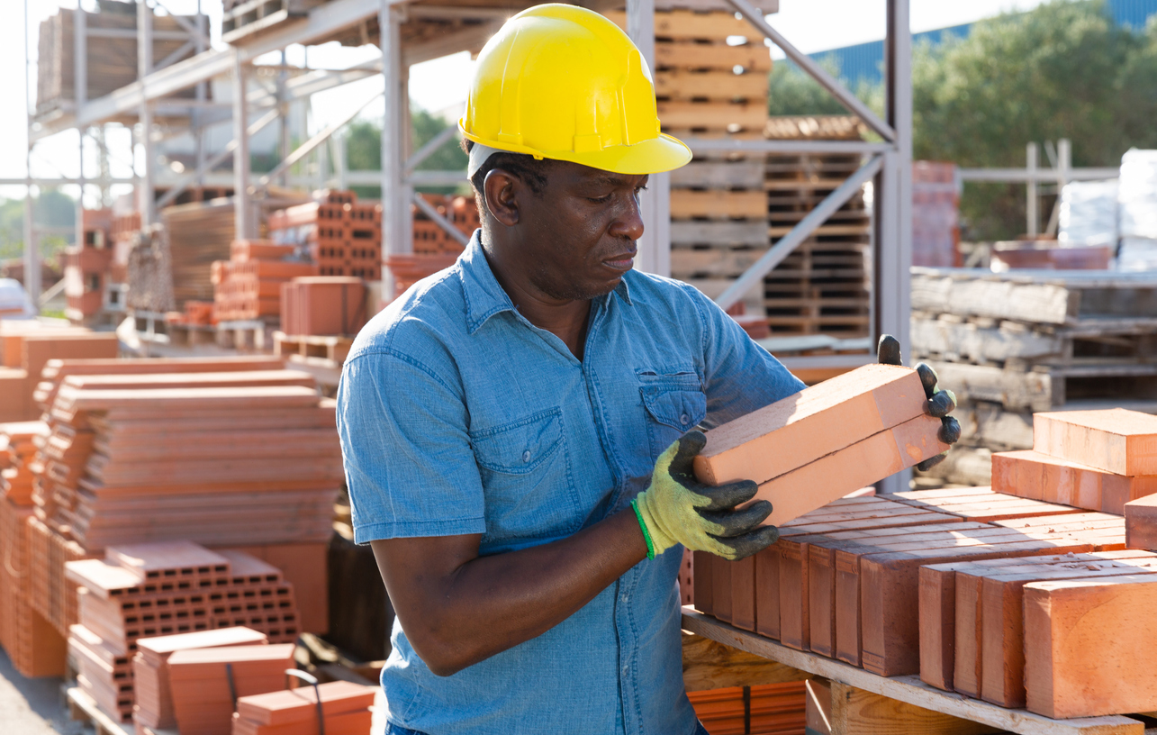African-american male worker carrying clay bricks in outdoor construction material storage.